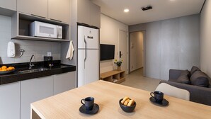 Una cocina moderna con microondas, refrigerador y lavamanos. Hay una mesa de comedor con dos tazas y un bol de pan. Se ve un sofá y un televisor al fondo.