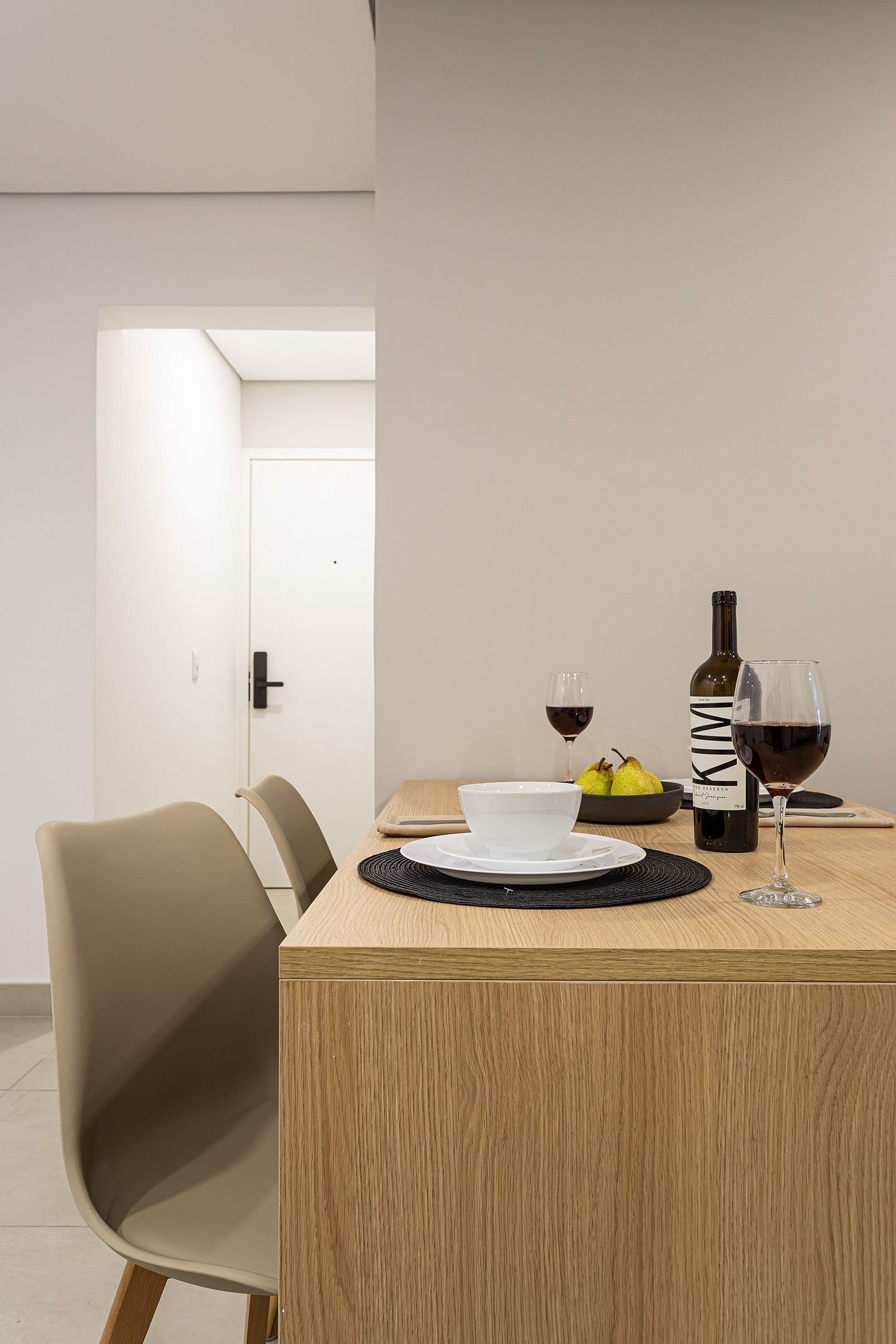 A dining area with a wooden table, two chairs, a bowl of fruit, a bottle of wine, and a wine glass.