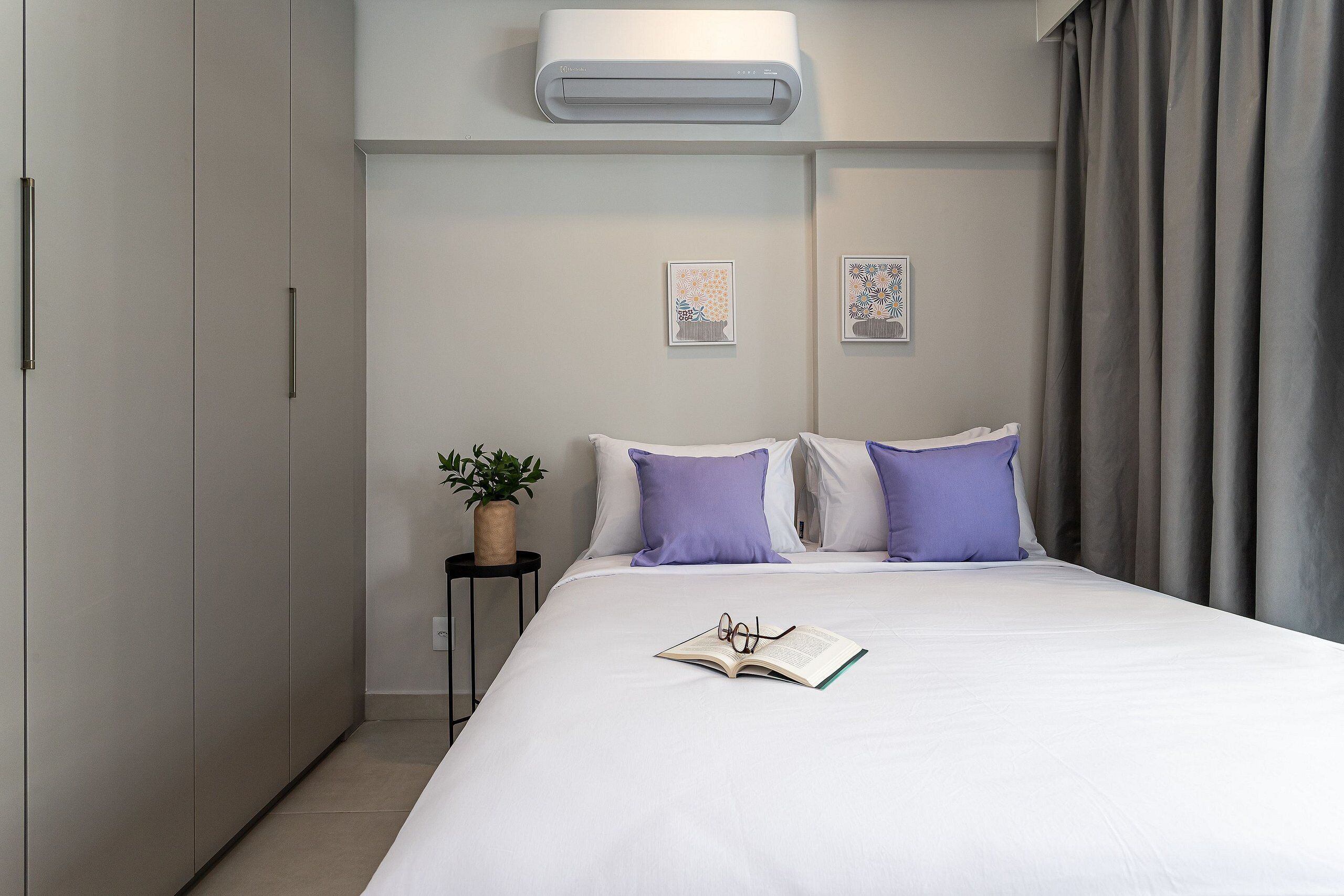 A modern bedroom with a large bed, a bedside table with a plant, and a wall-mounted air conditioner.