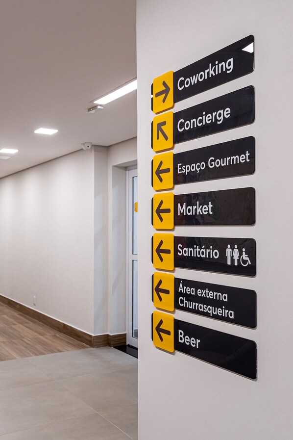 Parede com placas de sinalização indicando diversas comodidades: Coworking, Concierge, Espaço Gourmet, Mercado, Sanitário, área externa com churrasqueira e cerveja.