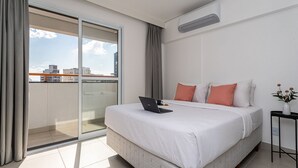 Habitación de hotel moderna con una cama grande, una computadora portátil sobre la cama, una mesita con un florero, y vista a la ciudad a través de una puerta corrediza de vidrio.