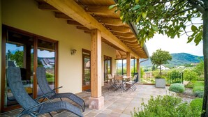 A covered patio with two lounge chairs, a table, and chairs, overlooking a landscaped garden and a mountainous landscape. - Beautiful vacation apartment on the edge of the Palatinate Mountains (Edenkoben)