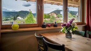 A dining area with a wooden table and chairs, a window with a view of a rural landscape, and a vase with flowers on the windowsill. - Beautiful vacation apartment on the edge of the Palatinate Mountains (Edenkoben)