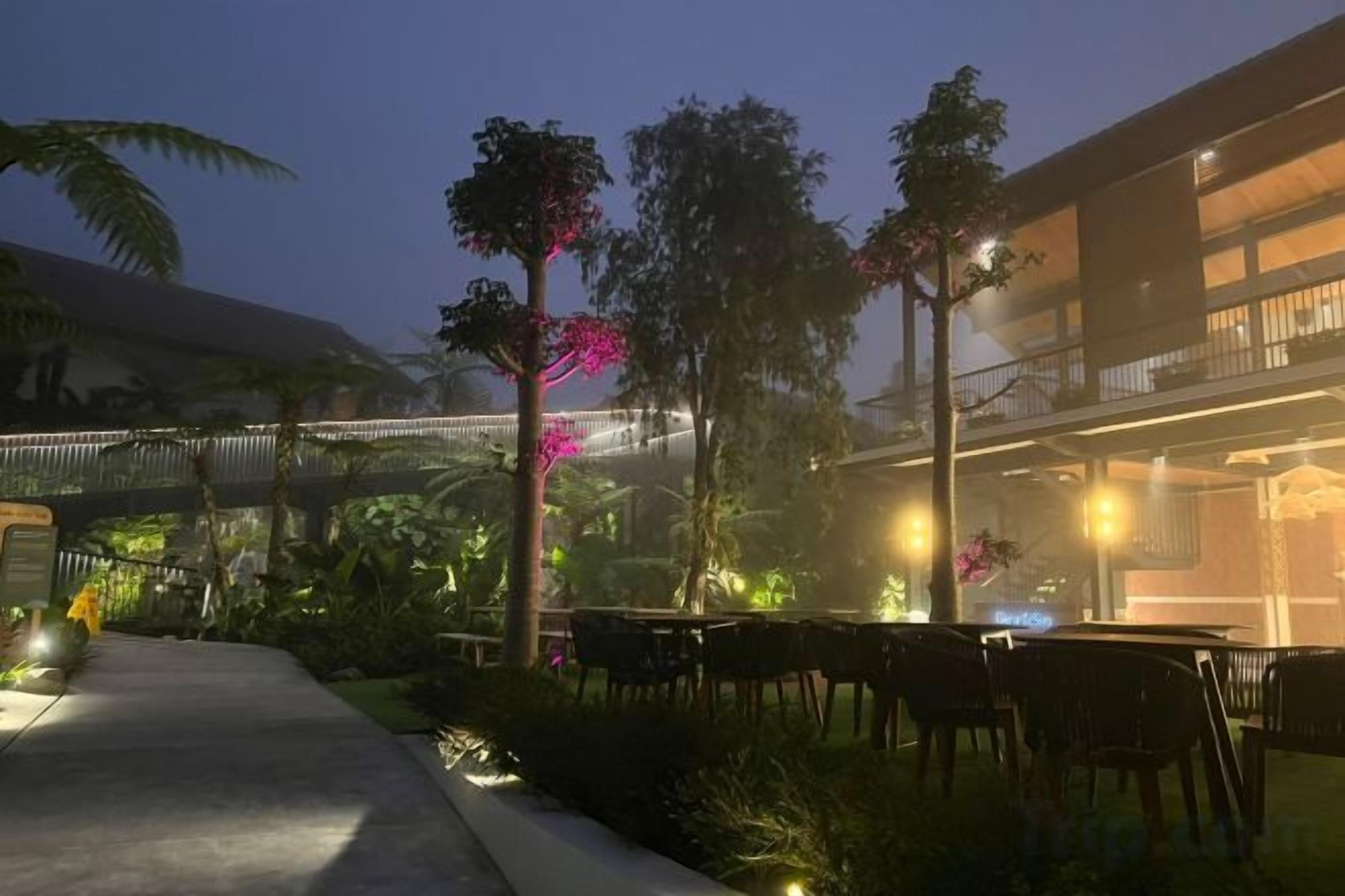 Un área de comedor al aire libre con mesas y sillas, rodeada de follaje y un edificio de varios pisos.