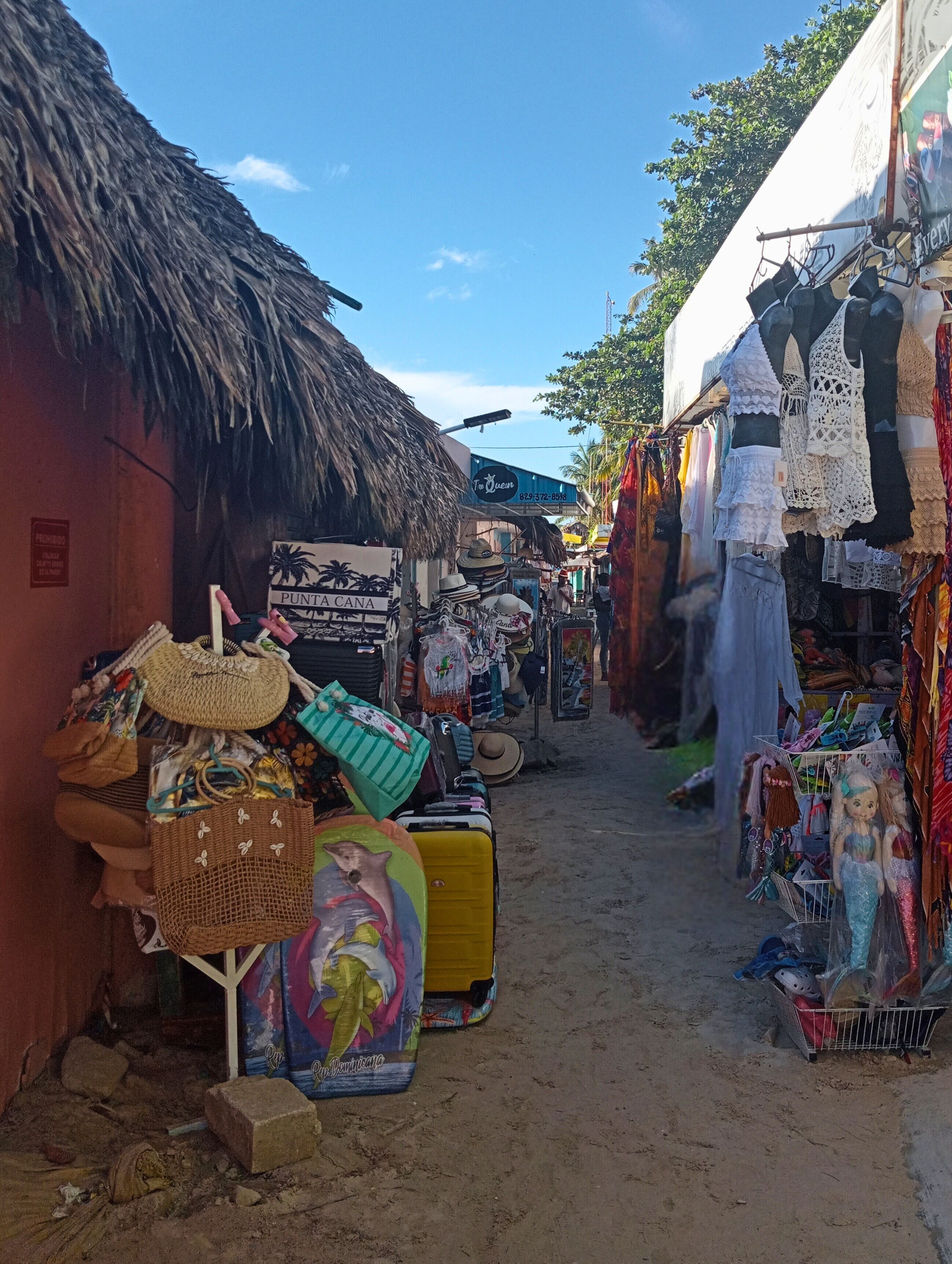 A narrow street with thatched-roof buildings and various stalls selling bags, hats, and other items.