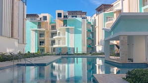 A swimming pool surrounded by multi-story buildings with balconies, a clear sky, and a calm water surface. - Maple Beach Residences Punta Cana  (Punta Cana)