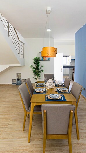 A dining area with a wooden table set for six, surrounded by grey upholstered chairs. A wooden pendant light hangs above the table, and a potted plant is on the floor beside it. - Maple Beach Residences Punta Cana  (Punta Cana)