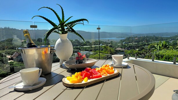 Een tafel met een schaal fruit, een fles, een vaas met een plant en twee kopjes op een balkon met uitzicht op een mooi panorama.