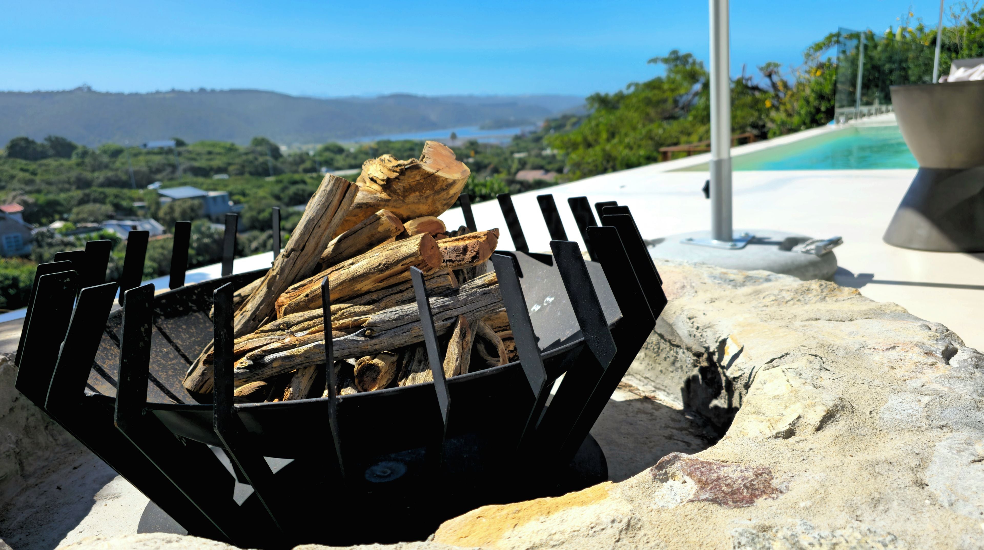 Un pozo de fuego negro con leña, rodeado de rocas, con una piscina y vegetación al fondo.