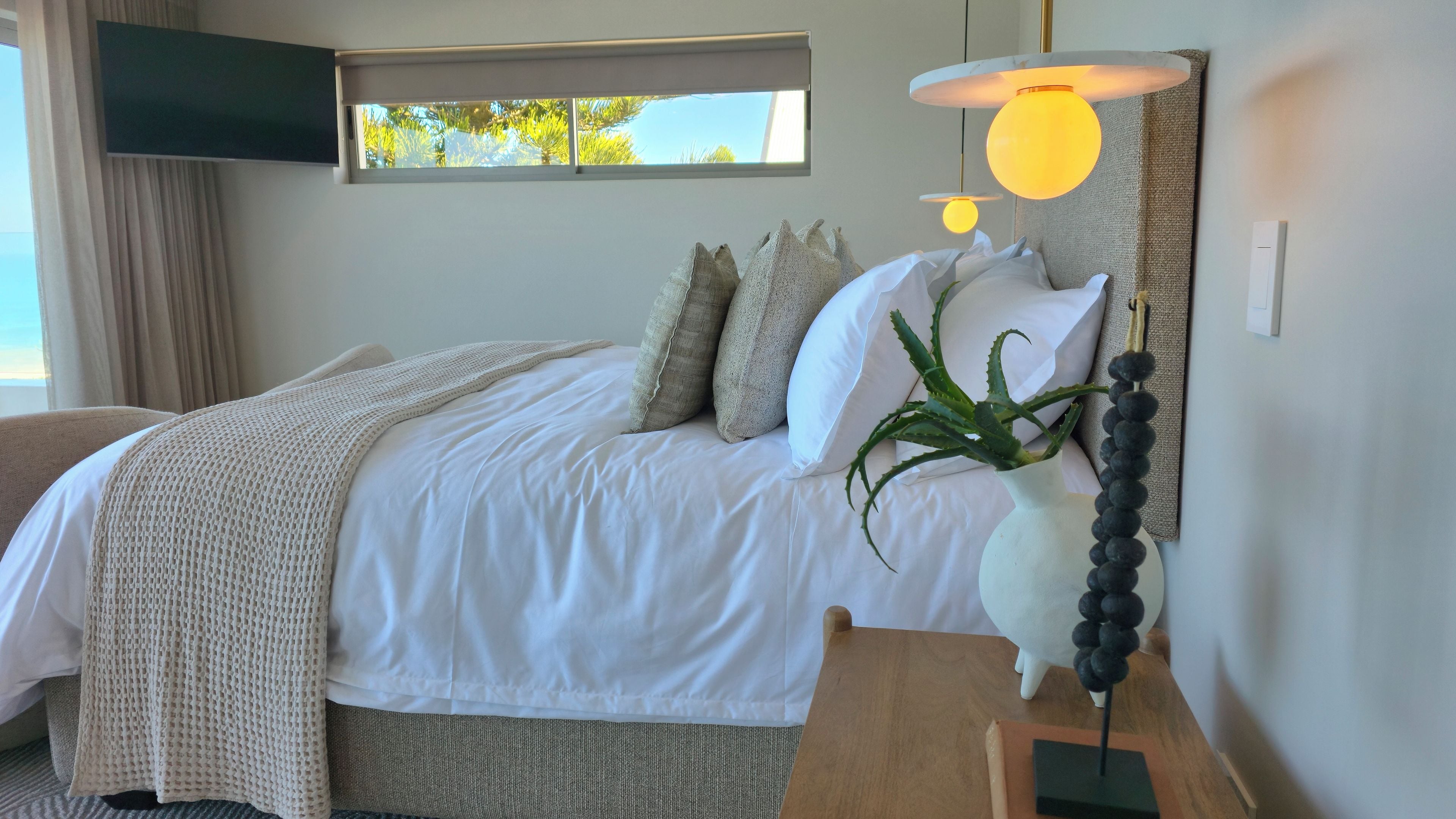 A neatly made bed with pillows and a blanket, a bedside table with a lamp, and a window with a view of trees.