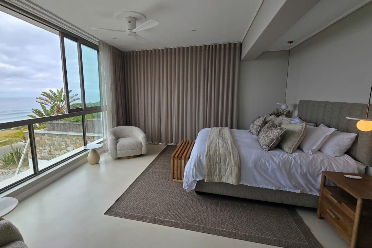 A modern bedroom with a large bed, a chair, and a view of the ocean.