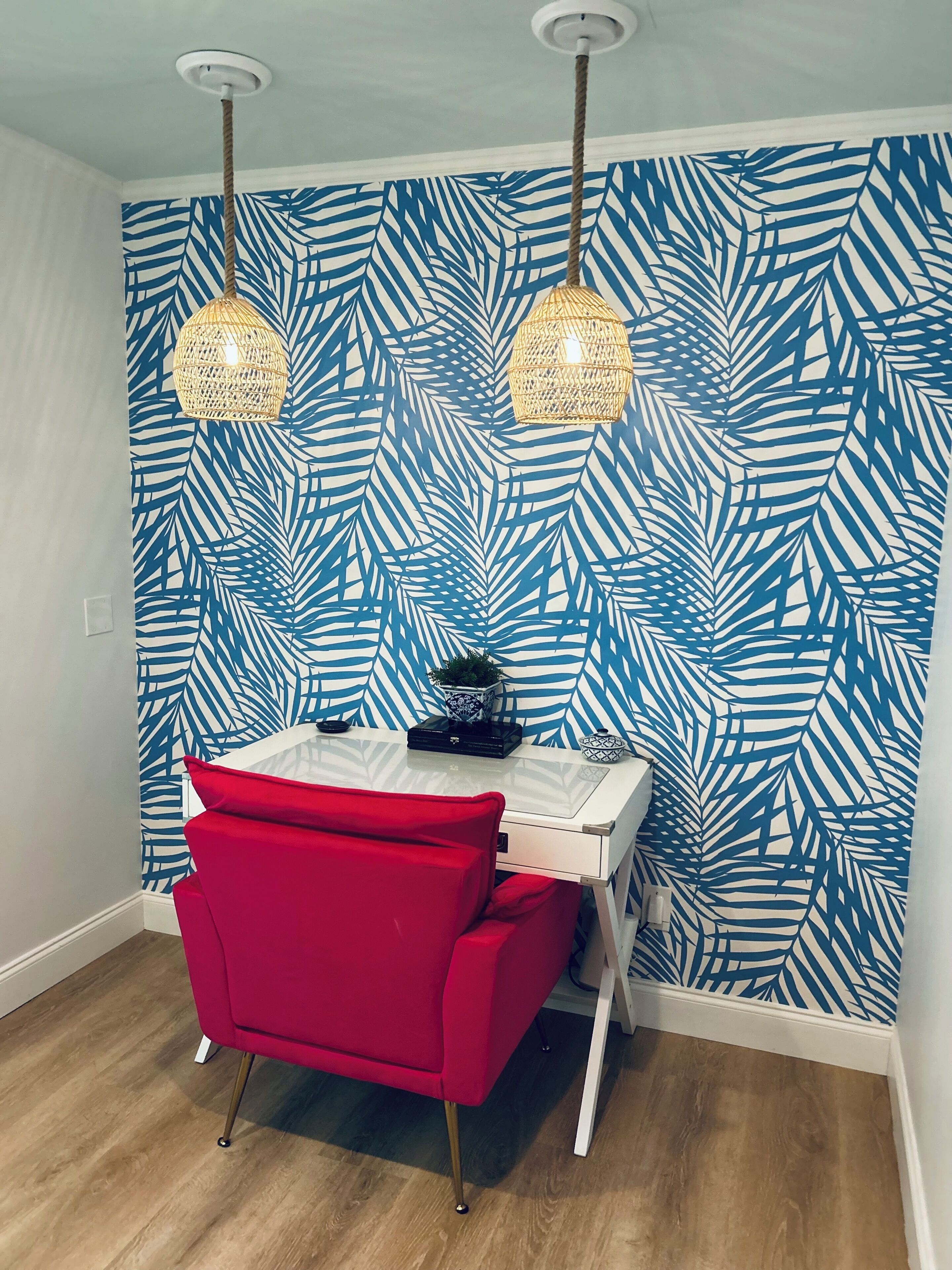 Ein Zimmer mit einem weißen Tisch, einem roten Stuhl und einer Wand mit blauem Blattmuster. Über dem Tisch befinden sich zwei Hängelampen.