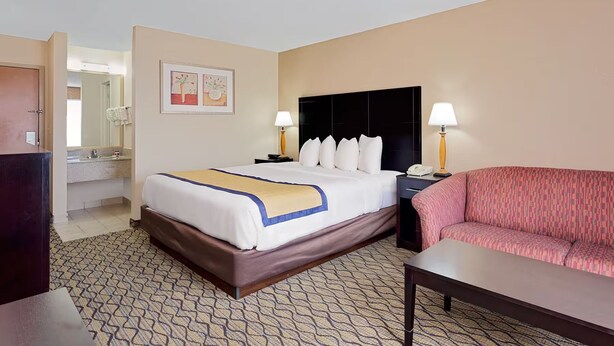 A hotel room with a bed, bedside lamps, a sofa, a desk, and a bathroom visible in the background. - Staywell Inn (Kalamazoo)