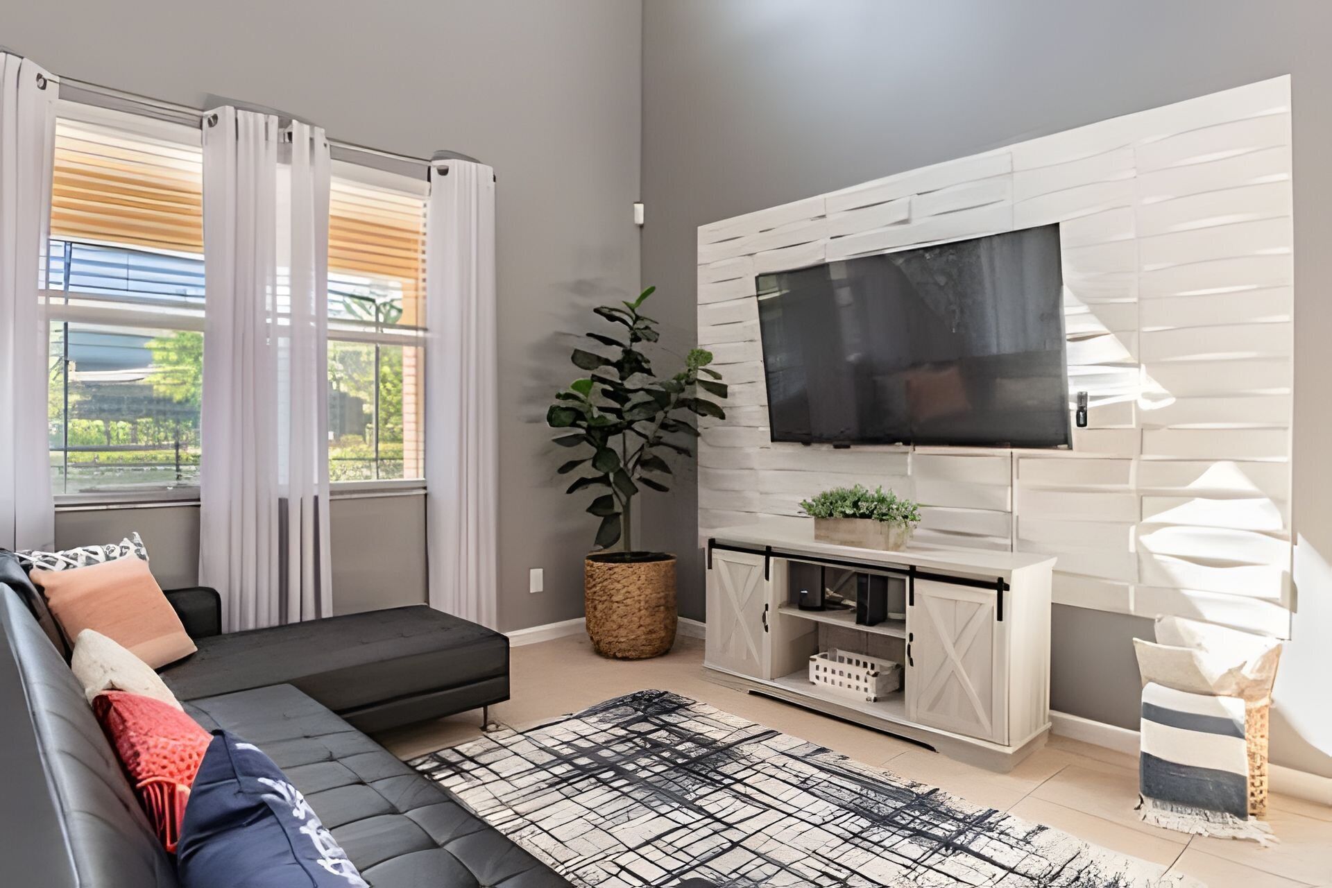 A modern living room with a black sofa, a flat-screen TV mounted on a white wall, a potted plant, and a large window with blinds.