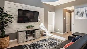 Un salon moderne avec un téléviseur à écran plat fixé à un mur blanc, un canapé gris, un tapis noir et blanc et une plante en pot.
