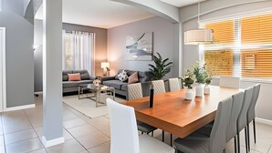 Un salon moderne avec un coin repas, comprenant une table à manger en bois, des canapés gris et une grande fenêtre avec des stores.