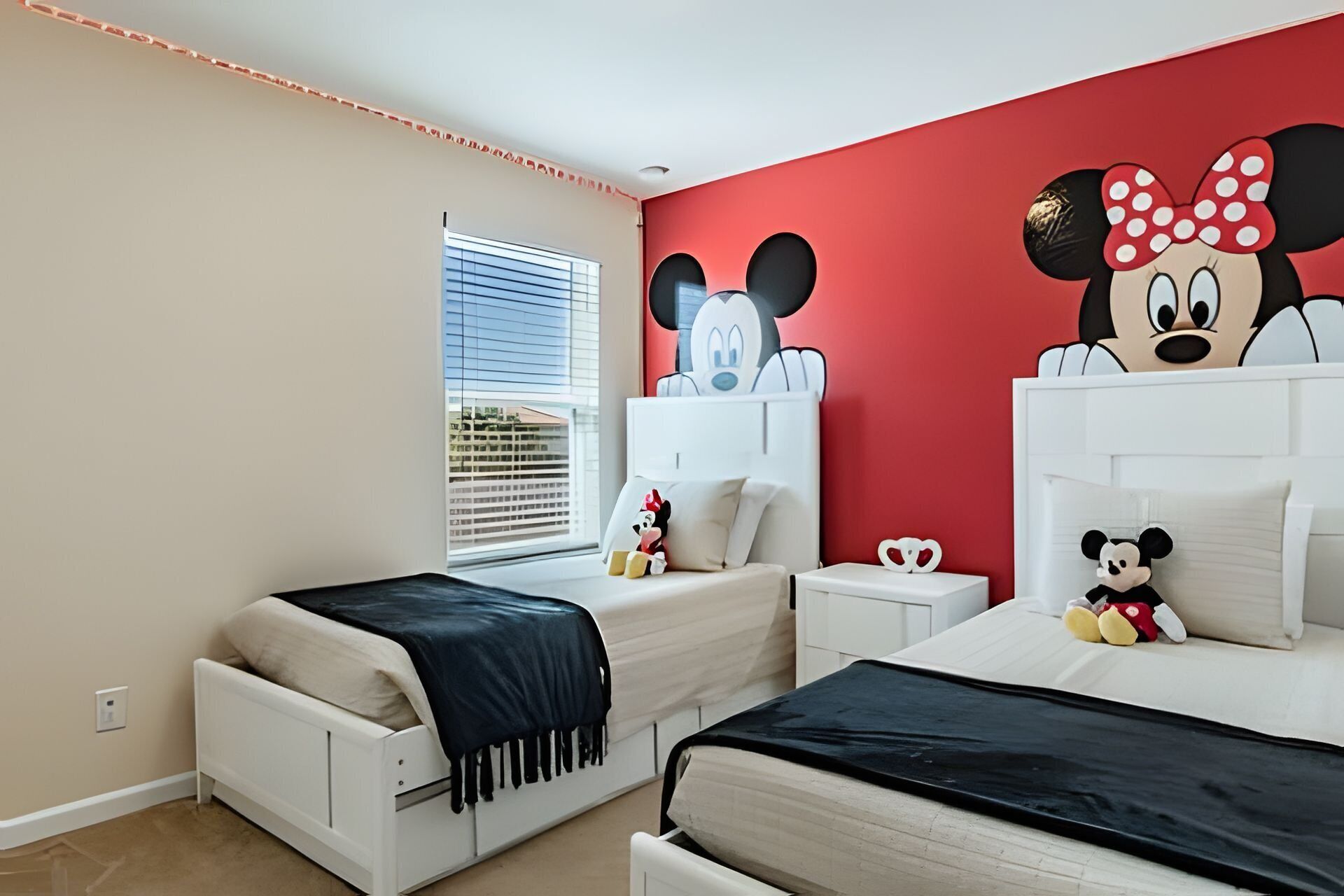 Habitación de hotel con dos camas, pared roja decorada con murales de personajes de dibujos animados, ventana con persianas y una mesita de noche con lámpara.
