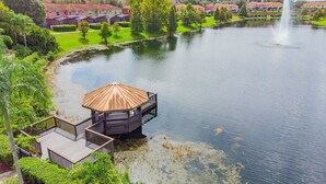 Ein See-Pavillon mit Holzterrasse, umgeben von üppiger Vegetation und freiem Blick auf eine Wohngegend im Hintergrund.