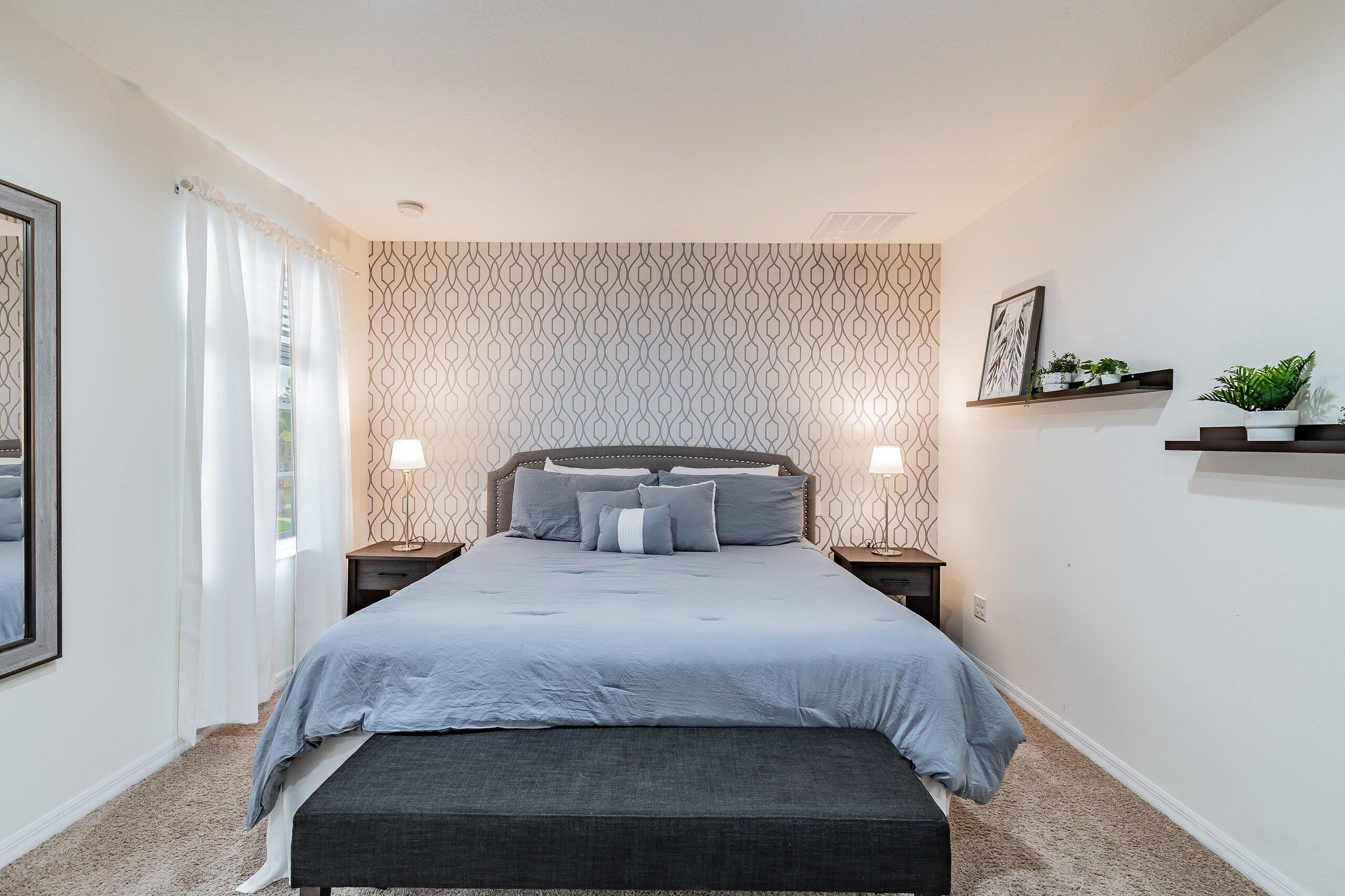 A bedroom with a bed, bedside tables, a bench, a window with curtains, and wall-mounted shelves with plants.