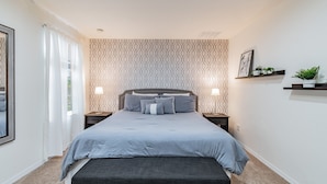 Ein Schlafzimmer mit einem Bett, Nachttischen, einer Bank, einem Fenster mit Vorhängen und Wandregalen mit Pflanzen.