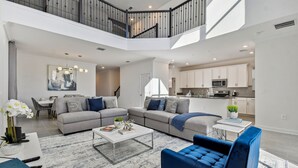 Amplia sala de estar con un sofá gris, un sillón azul y una mesa de centro blanca. Una escalera conduce a un nivel superior con balcón.