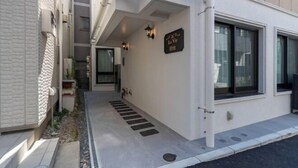 A narrow pathway leading to a building entrance with a sign above the door, a small outdoor seating area, and a window with a view. - LaVie Hatsudai (Tokyo)