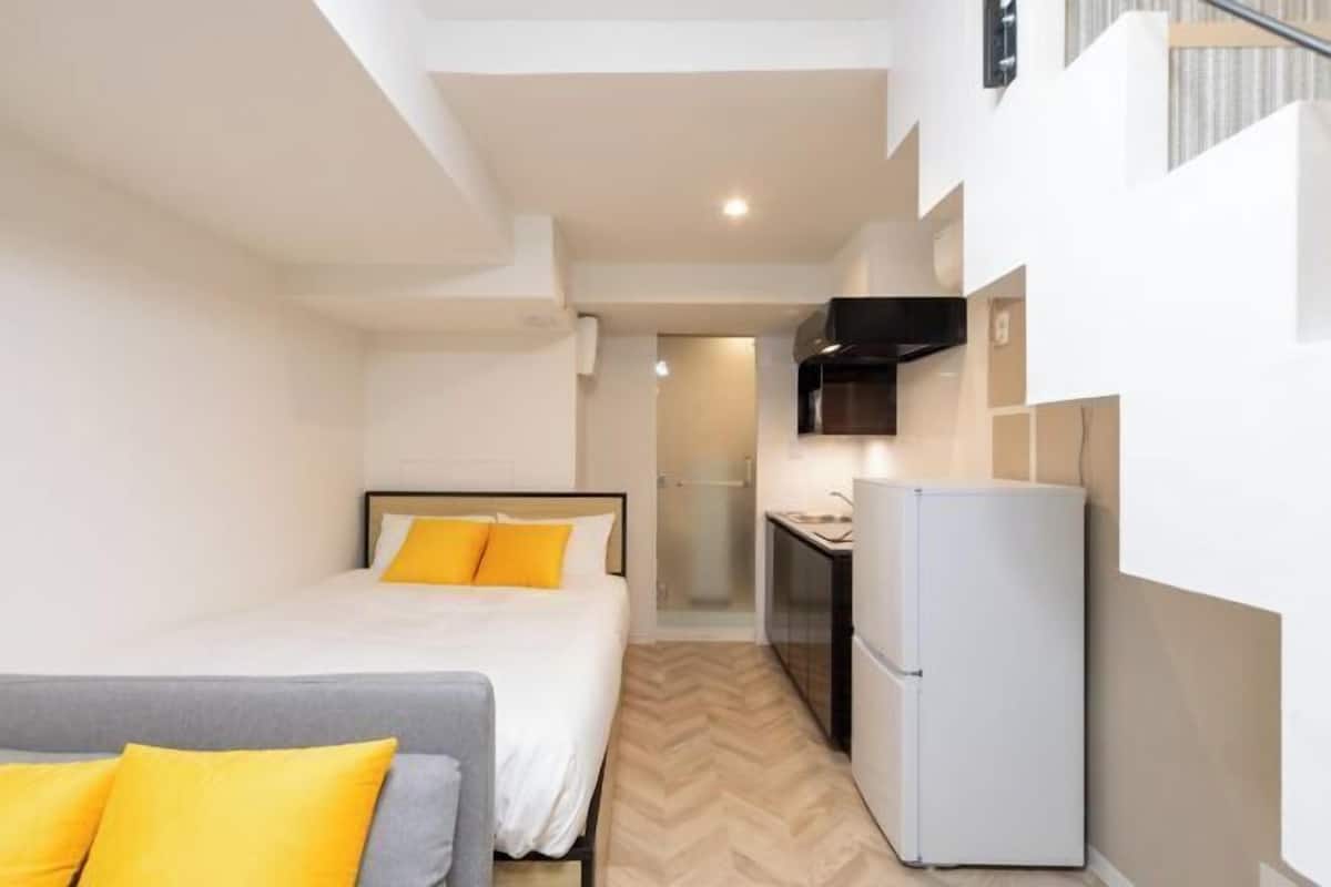 A compact hotel room with a bed, a sofa, a kitchenette, and a staircase.
