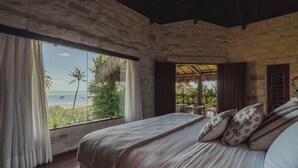 A bedroom with a stone wall, a large bed with patterned pillows, a view of the beach, and a thatched roof structure outside.