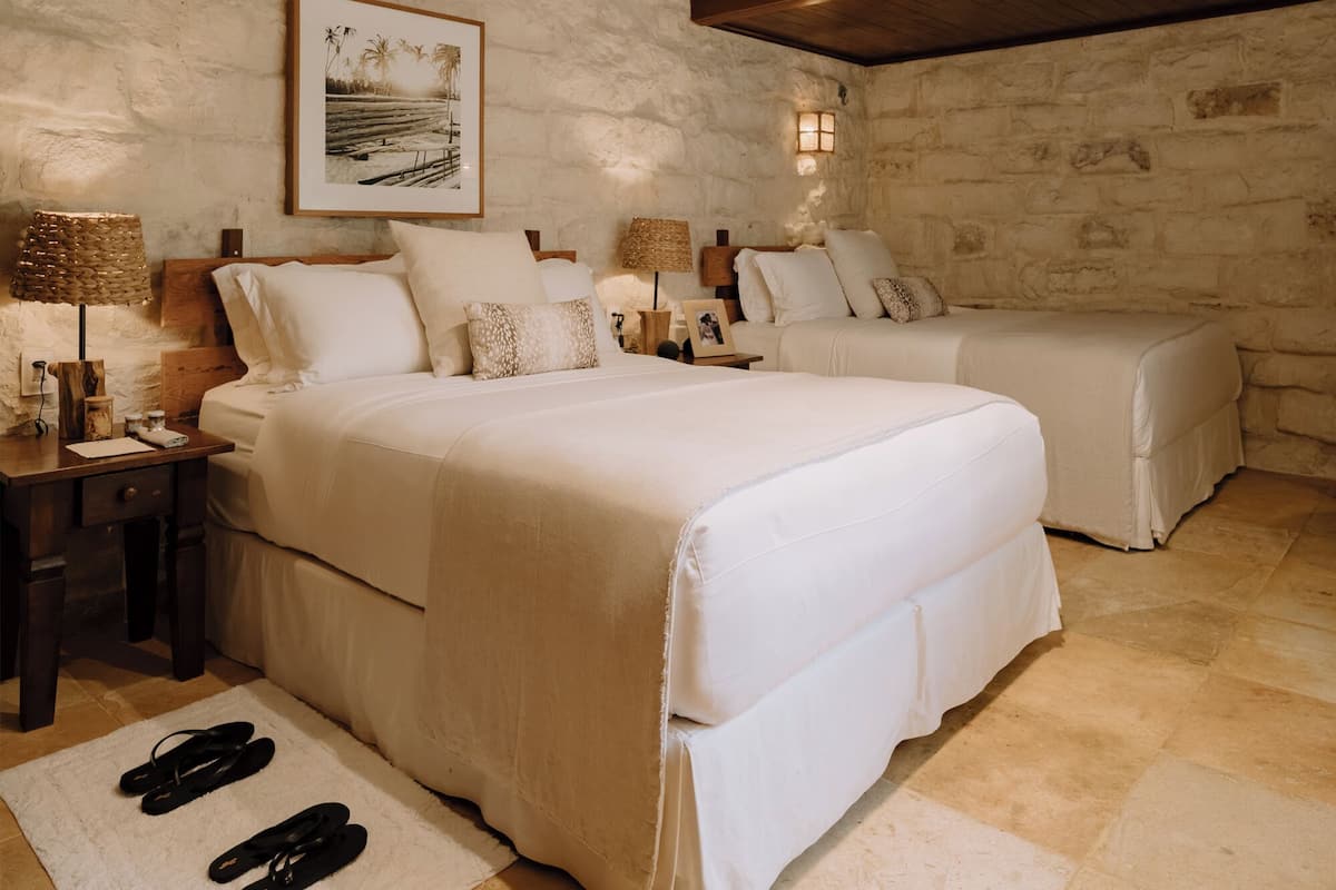 Une chambre avec un mur en pierre, un lit avec une literie blanche, deux tables de chevet avec des lampes et un tableau encadré au mur.