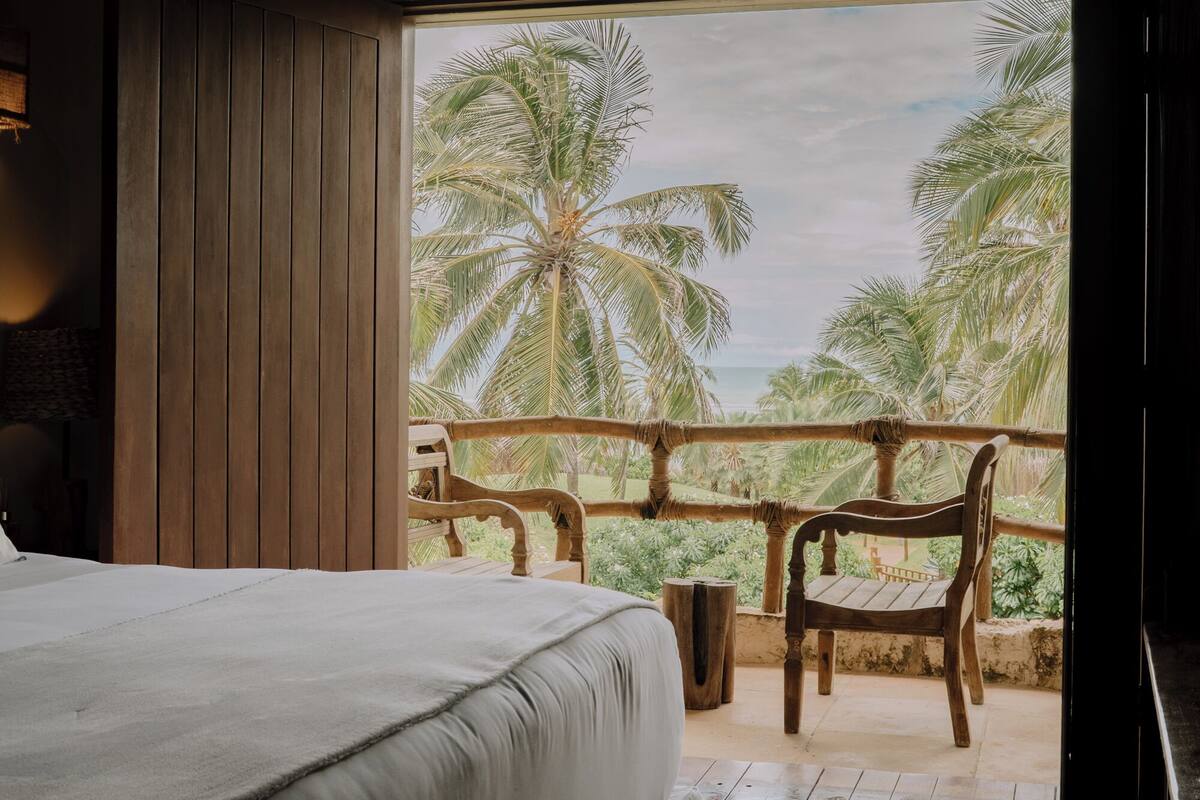Une chambre avec un grand lit, des meubles en bois et un balcon offrant une vue sur les palmiers et la mer.