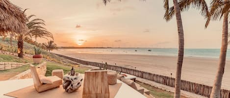 Un salón junto a la playa con mobiliario de madera y vistas al atardecer sobre el océano.