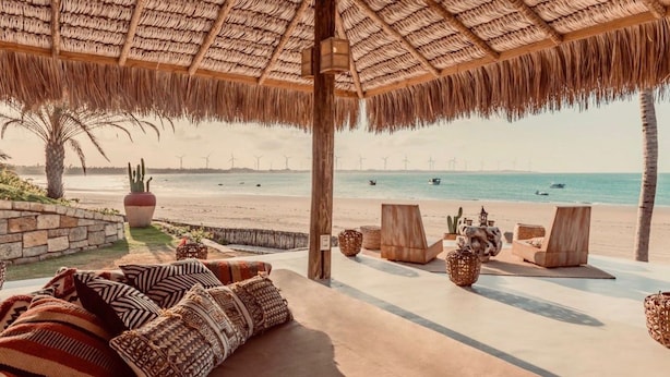 A beachside lounge with a thatched roof, wooden furniture, and a view of the sea and wind turbines.