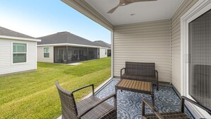 A covered patio with wicker furniture and a ceiling fan, overlooking a grassy yard and neighboring houses. - Spacious 2BR in The Villages | GolfCart | King Bed (Sumterville)