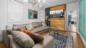 Un salon moderne avec un canapé d’angle, une table basse en verre et un tableau représentant un paysage urbain accroché au mur.