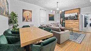 Un salon moderne avec une table à manger en bois, des chaises recouvertes d’un tissu vert, un canapé gris et une œuvre d’art murale représentant un paysage urbain.