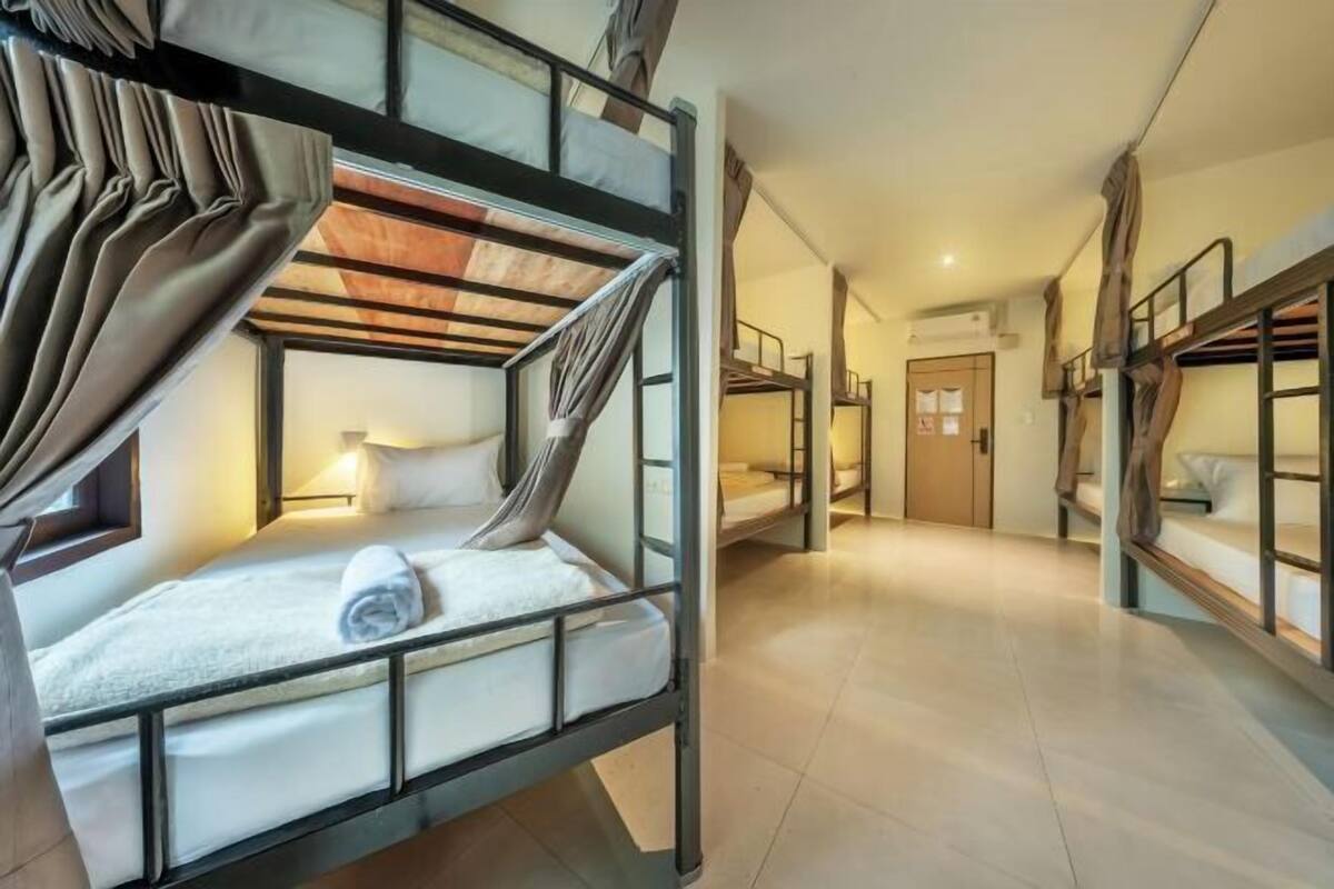 A dormitory room with bunk beds, a window with curtains, a bedside table, and a lamp.
