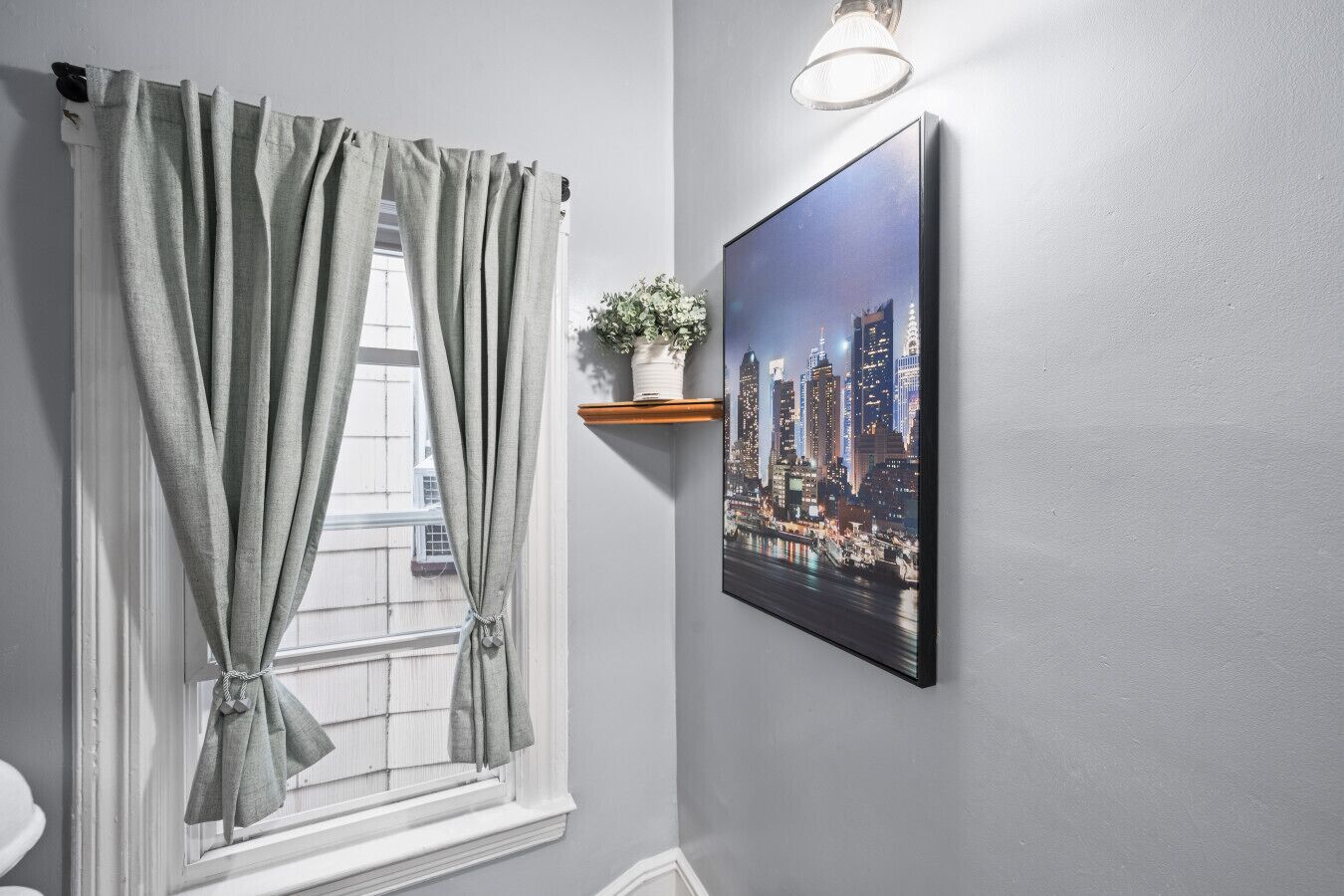 Une salle de bain avec une fenêtre, des rideaux gris, une plante en pot posée sur une étagère et un tableau représentant un paysage urbain accroché au mur.
