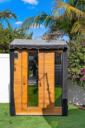 Una pequeña estructura de madera con techo negro, una puerta y una ventana, rodeada de hierba verde y plantas.