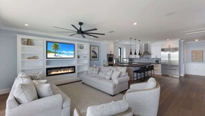 A modern living room with a fireplace, a large TV, and a kitchen area with a stainless steel refrigerator. - Aqua - Brand New Luxury Beach House on AMI (Bradenton Beach)