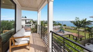 A balcony with a wooden ceiling, a white sofa, and a view of a playground and water body. - Aqua - Brand New Luxury Beach House on AMI (Bradenton Beach)
