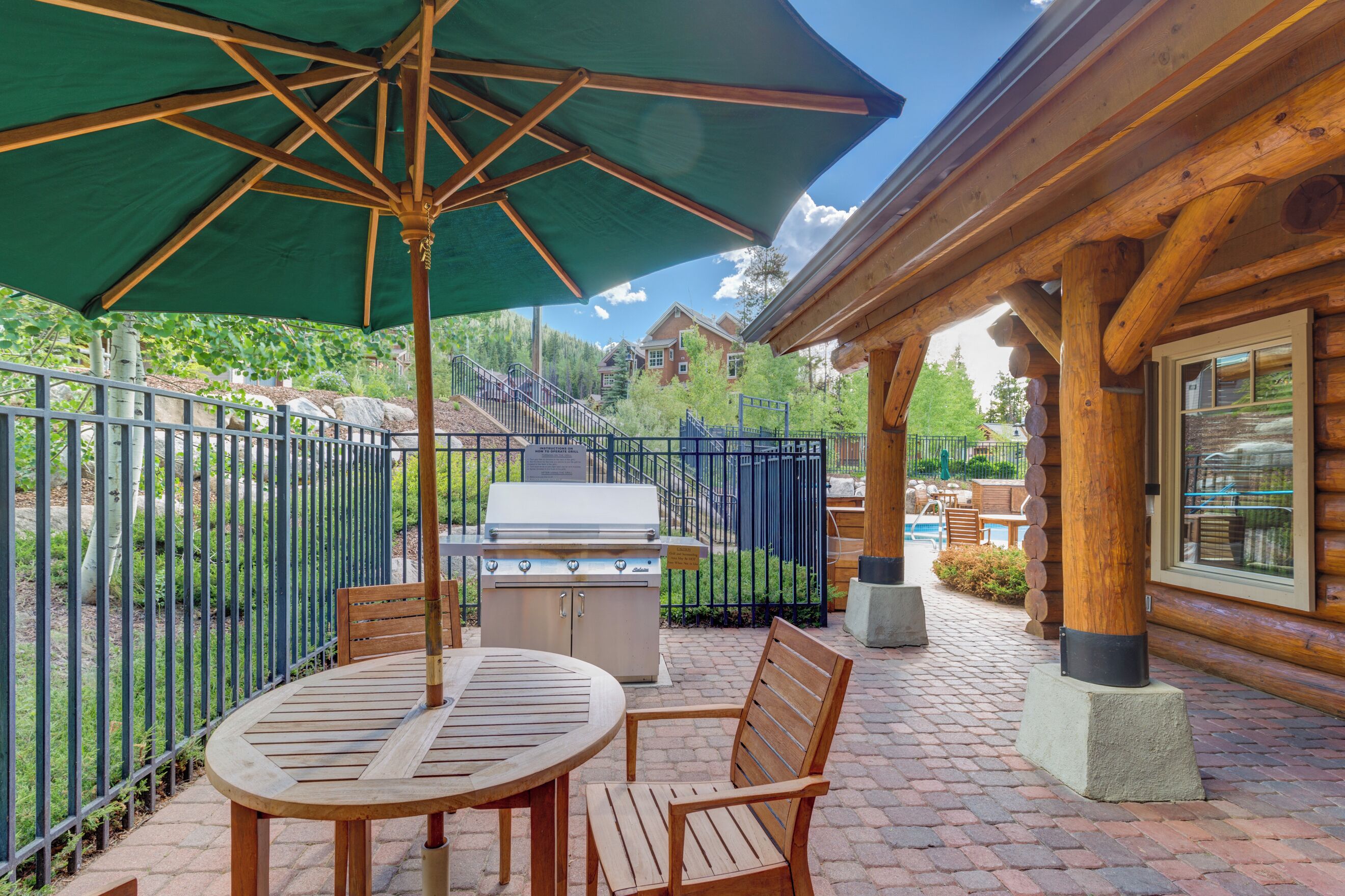 An outdoor patio with a grill, wooden chairs, and a table under a green umbrella.