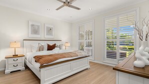 Une chambre à coucher comprenant un lit, des tables de chevet, un ventilateur de plafond et de grandes fenêtres équipées de volets.