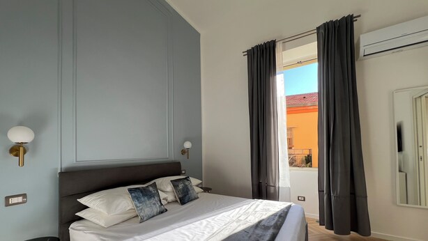 A neatly made bed with white linens and a dark headboard, a small bedside table with a lamp, and a view of an outdoor balcony through a window with dark curtains. - Comfortable and stylish room near central station and airport (Napoli)