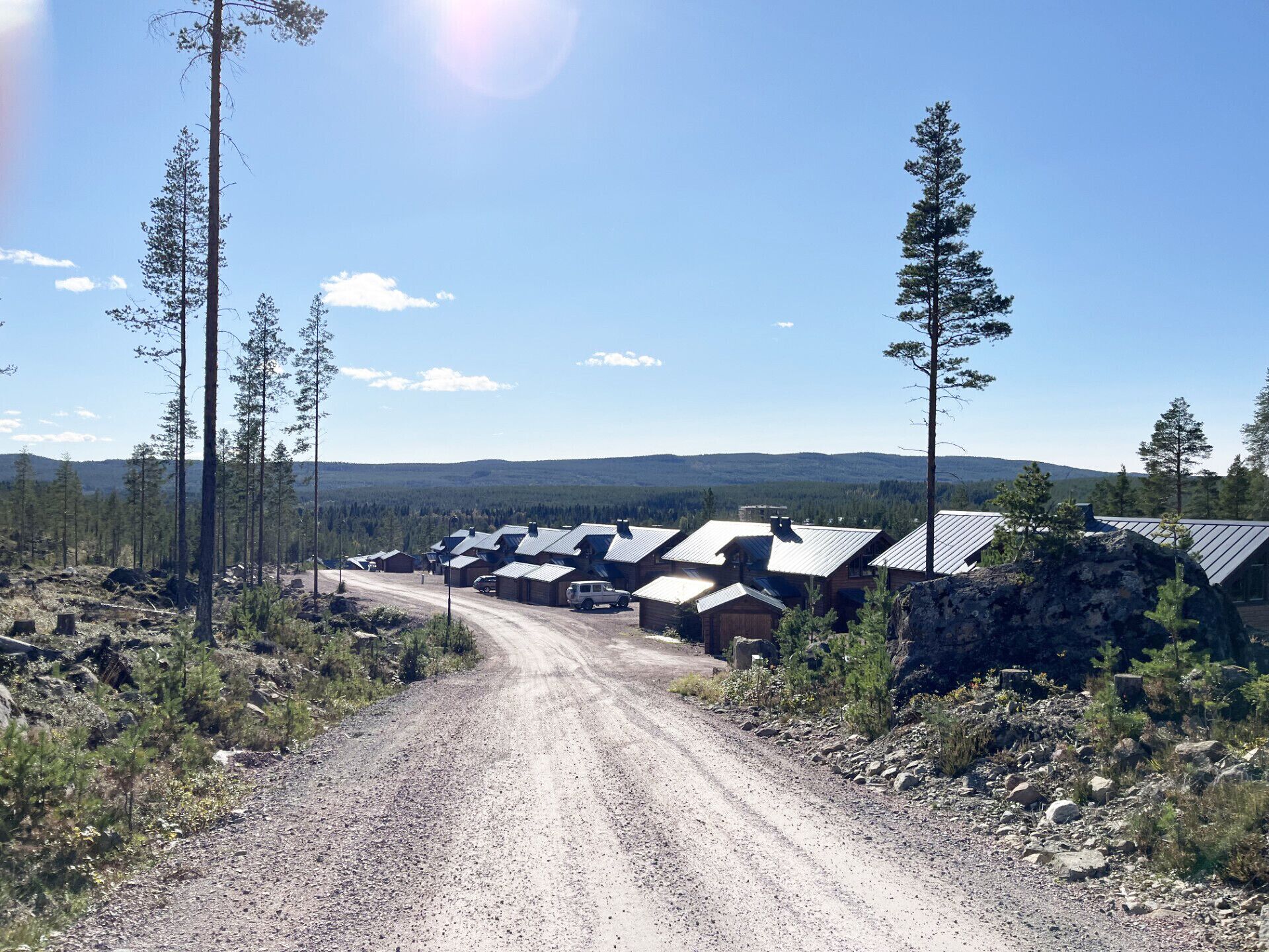 A gravel road leading to a row of cabins with metal roofs, surrounded by trees and a mountainous backdrop.