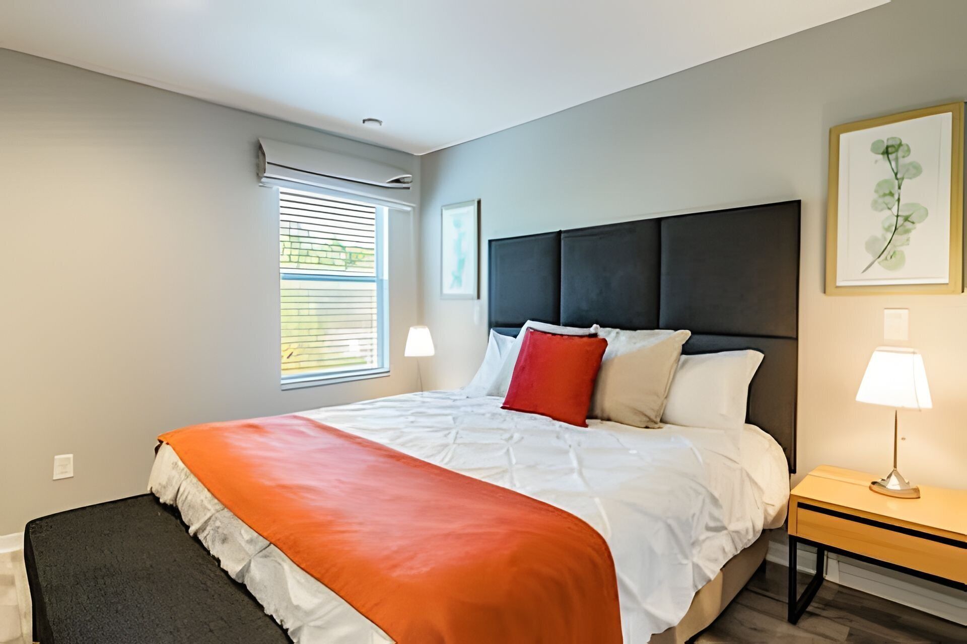 A modern bedroom with a large bed, a nightstand, a window with blinds, and a framed picture on the wall.