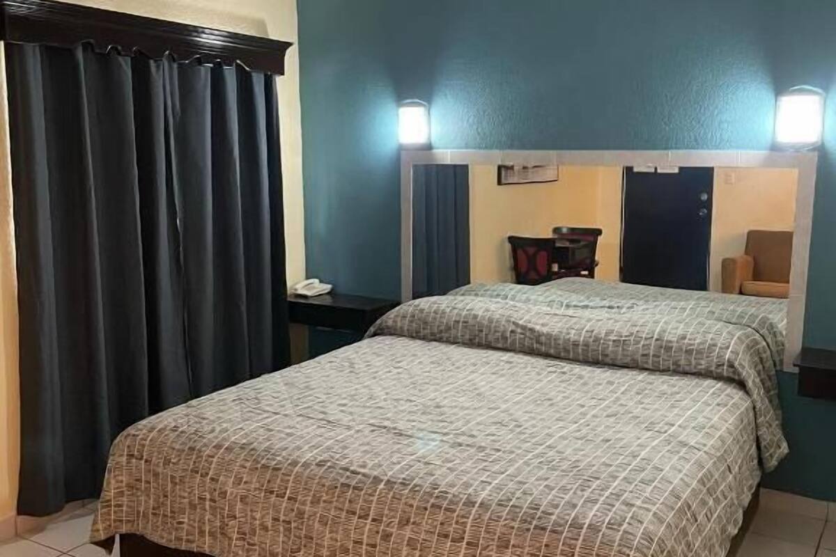 Une chambre d’hôtel avec un lit, un ventilateur de plafond, une table de chevet, un miroir et une fenêtre avec des rideaux.