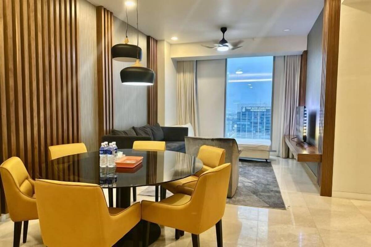 A modern hotel room with a dining area featuring a round table and yellow chairs, a sofa, and a city view.