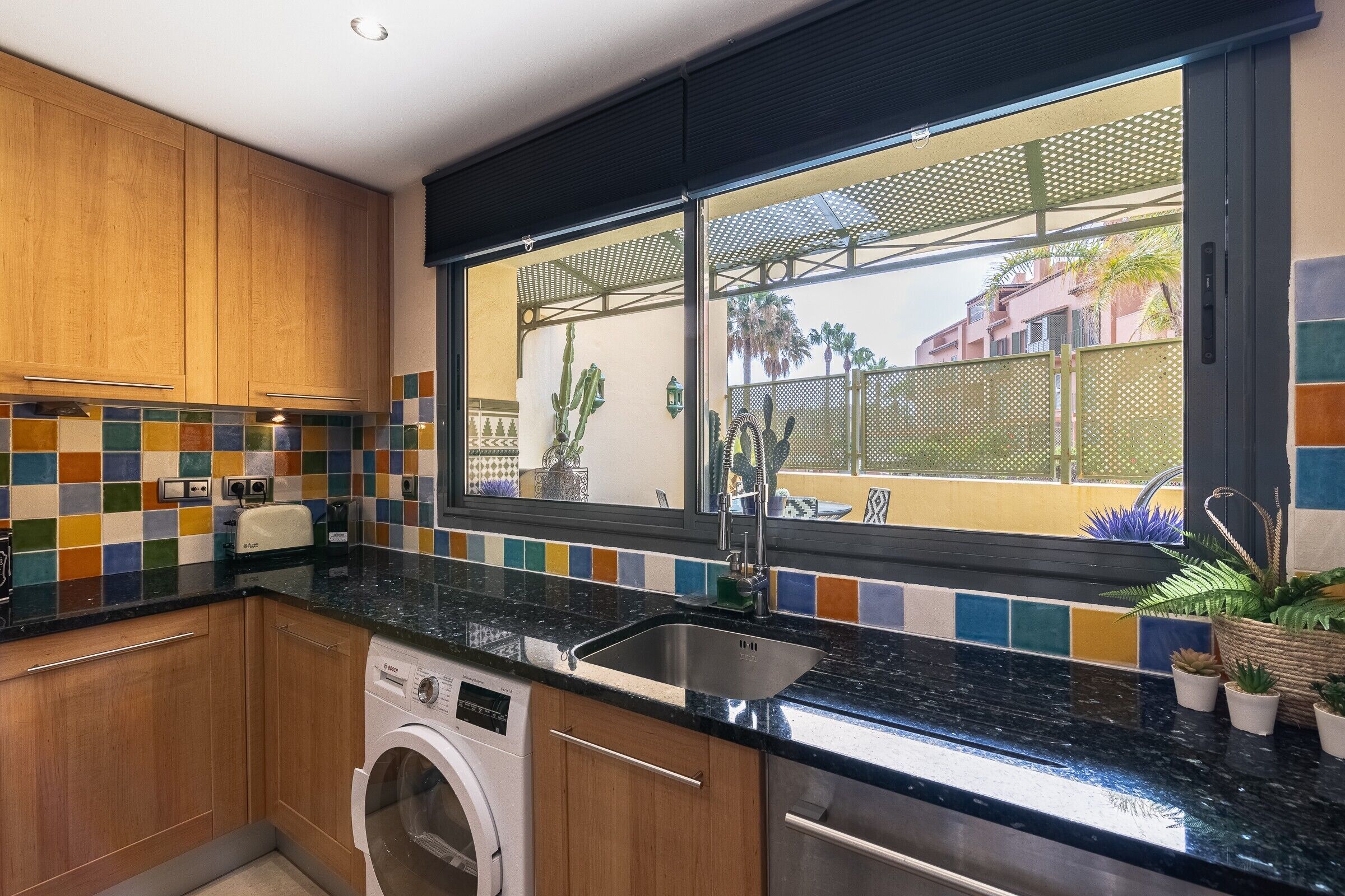 Een keuken met houten kastjes, een wasmachine, een spoelbak en een raam met uitzicht op palmbomen en gebouwen.
