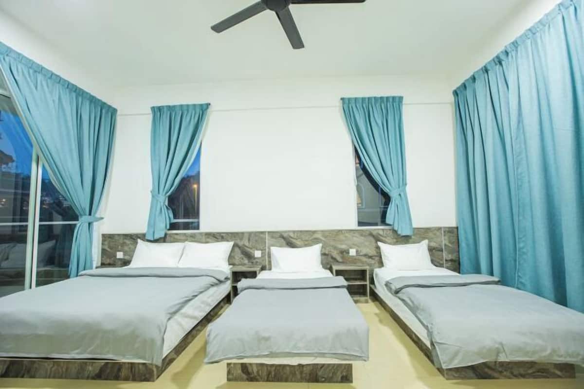 A hotel room with three single beds, a ceiling fan, and large windows with blue curtains.