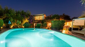 En swimmingpool med en klar, blå vandoverflade, omgivet af frodigt grønt og en oplyst bygning i baggrunden.